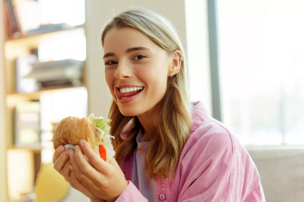 mujer mostrando los dientes mientras sostiene una hamburguesa, una manera de explicar la función de los dientes en la masticación, habla y estética facial.